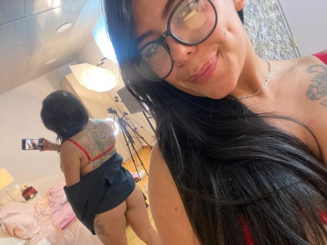 CatalinaReiyna, usando óculos, com cabelo preto longo, sutiã vermelho, saia preta puxada para cima, mostrando sua bunda e tatuagens nas costas, sorrindo, tirando uma selfie em um quarto..
