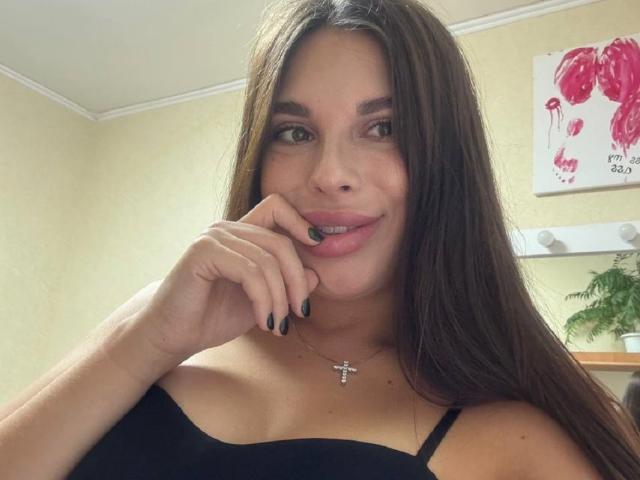 BlackFatal con cabello castaño largo, uñas negras, top con tirantes negros, ligera sonrisa, mano cerca de la boca, collar con cruz negra, piel morena, ojos marrones, iluminación suave, linda, atractiva..