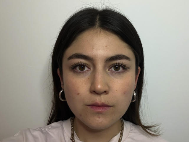 SweetSella has brown eyes, dark straight hair, and wears hoop earrings with a white shirt. Her face shows acne and she has a slight pout.