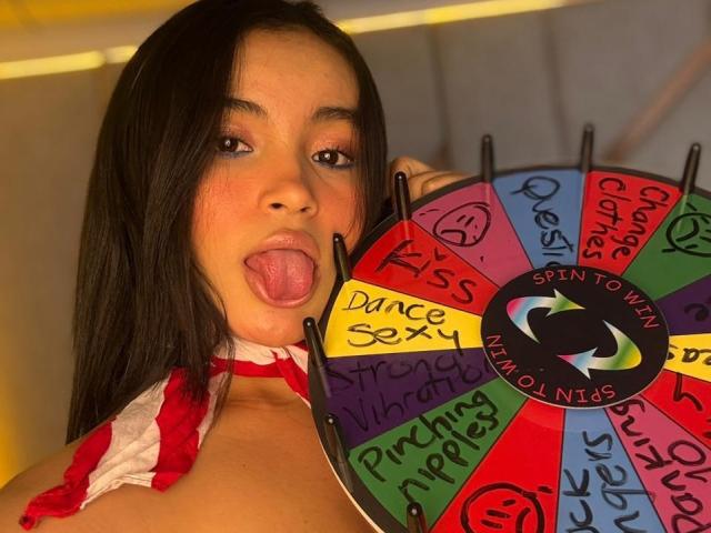 MiiaPalmer, avec ses longs cheveux noirs, sa langue tirée, tenant un jeu de toupie coloré, portant un haut à bretelles rouge et blanc, montrant ses seins nus..
