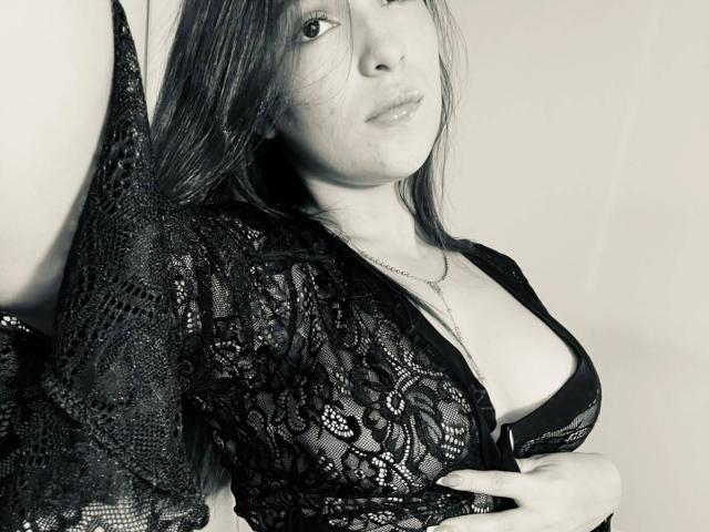 Katarina Novak, avec de longs cheveux, portant une robe en dentelle noire, laissant entrevoir ses seins dans un soutien-gorge noir, lèvres légèrement entrouvertes, regardant la caméra, une main sur la robe. Monochrome..