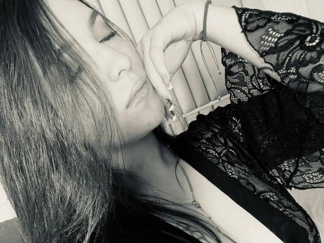 Katarina Novak, con lunghi capelli lisci, occhi chiusi, indossa una camicetta di pizzo nero, toccandosi la guancia con la mano destra, mostrando una collana d'argento e un braccialetto..