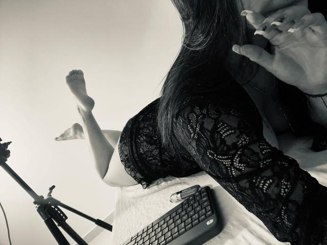 Katarina Novak liegt im Bett, die Beine hochgelegt, trägt ein schwarzes Spitzenkleid, eine Tastatur liegt auf dem Bett, ein Stativ ist links, die Hände sind in der Nähe des Gesichts, ein Schwarz-Weiß-Foto, der Fokus liegt auf der Spitze, das nackte Gesäß ist sichtbar..