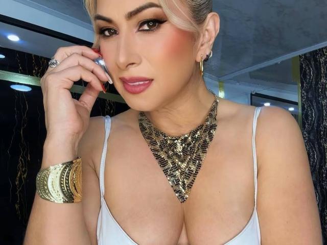 MisseScarleth with blonde hair, pink lipstick, and bold eye makeup, wearing a gold necklace, white top, and gold bracelet, holding her phone to her ear. Boobs, cleavage.
