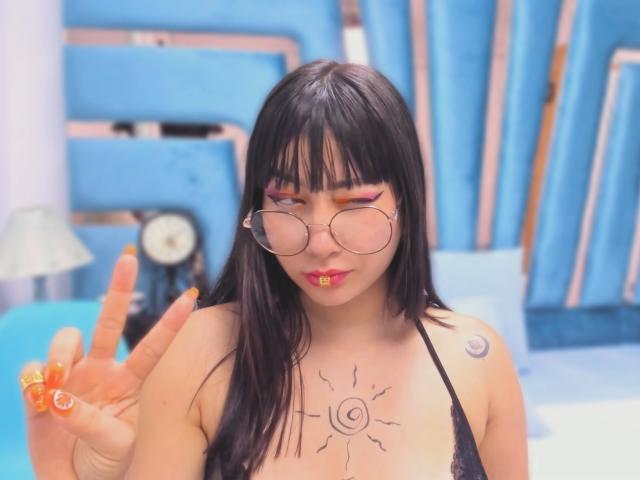 Genesiss with black hair, round glasses, and orange lipstick, making a peace sign with orange nails, has sun tattoo on chest and crescent moon tattoo on left shoulder.