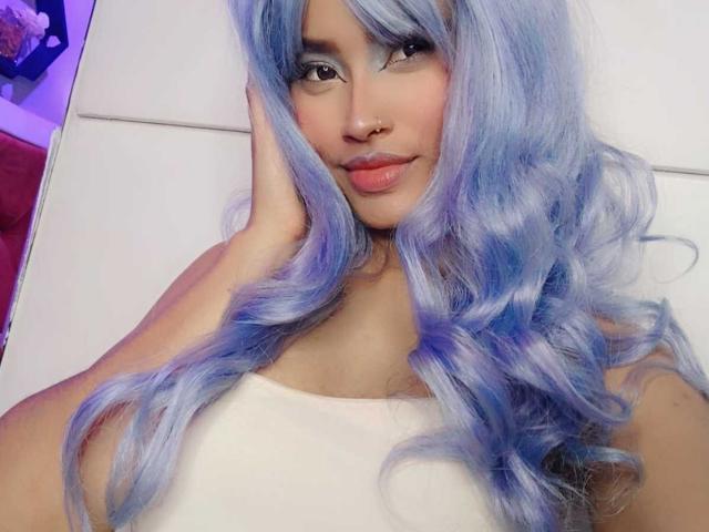 KarolPalacios, avec ses cheveux bouclés bleus, porte un haut blanc, a une main sur sa joue, sourit avec des lèvres roses, porte un anneau au nez et un maquillage léger. Les seins de KarolPalacios ne sont pas visibles..