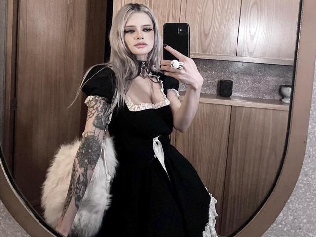 ErgoProxy com cabelo loiro comprido, vestido de empregada preto, babados brancos, braço tatuado, segurando um acessório branco fofo, tirando uma selfie no espelho, usando um colar preto, com expressão séria, segurando o celular..