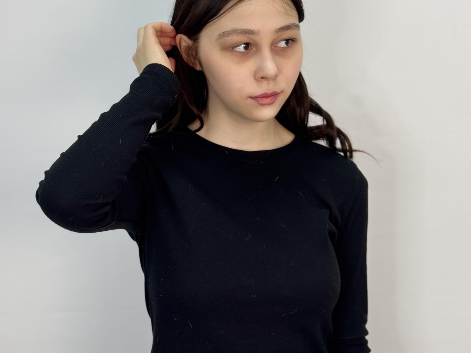 MaryaDottery, avec ses cheveux bruns, portant une chemise noire à manches longues, a une main dans ses cheveux, regarde légèrement sur le côté, avec une expression neutre, de petites seins et une peau pâle..