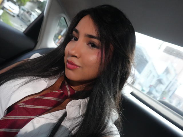 Melanie Cole mit langem, schwarzem Haar, die ein weißes Hemd und eine rot gestreifte Krawatte trägt, sitzt in einem Auto. Sie hat rosafarbenen Lippenstift und dezentes Make-up und blickt leicht zur Seite..