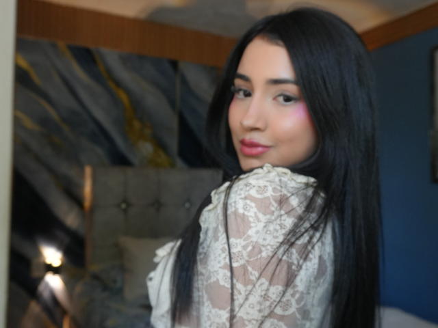 Melanie Cole, met lang zwart haar, roze oogschaduw, een witte kanten top, kijkt om en glimlacht, terwijl ze haar achterwerk laat zien. Zachte belichting, licht onscherpe achtergrond..