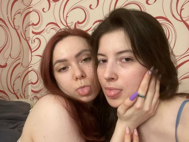 LeonoraGrego met rood haar en LeonoraGrego met zwart haar, beiden steken hun tongen uit, LeonoraGrego's hand op de wang van de LeonoraGrego met zwart haar, paarse ring, in een slaapkamer..