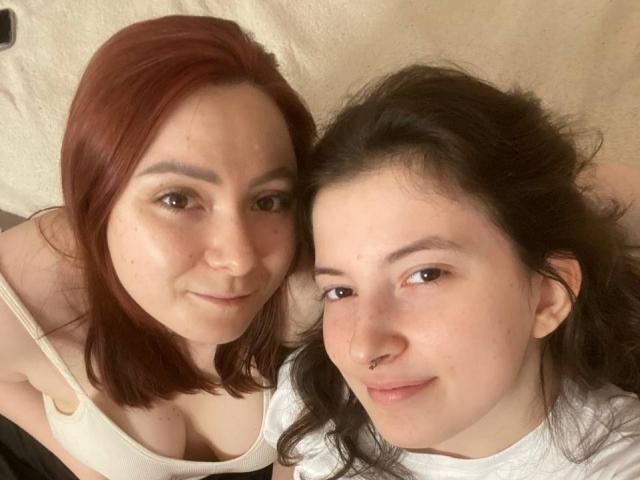 LeonoraGrego met rood haar en LeonoraGrego met bruin haar, beiden poserend in een selfie waarbij hun borsten zichtbaar zijn, glimlachend naar de camera, liggend, met zichtbare neus piercings..