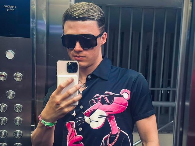 ErosHot, avec ses cheveux bruns lissés en arrière, ses lunettes de soleil noires et son t-shirt avec un léopard rose, prend un selfie dans un ascenseur, tout en portant un bracelet vert..