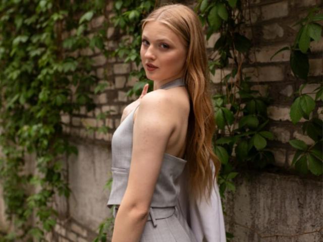 VandaMange met lang, golvend blond haar, die een grijze top met een halterhals draagt, kijkt over haar schouder en laat haar kleine, ronde billen en een subtiele glimp van haar borst zien..