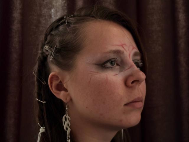 Johnetta Urtiaga, met een gezwollen gezicht, donker gevlochten haar, metalen kruisjes in haar haar en oorbellen met een ketting, kijkt opzij met een nadenkende blik, en vertoont rode krassen..
