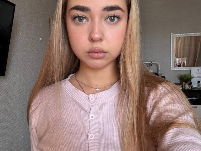 Georgine Branen, com cabelo loiro, olhos verdes e um suéter rosa, abotoado, olhando para a câmera com os lábios curvados. Seus seios são pequenos e ela tem um rosto bonito..