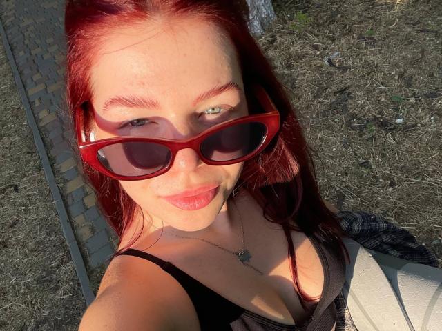 AlexXas, met rood haar, draagt rode zonnebril, een zwart tanktopje, toont haar borsten, heeft een lichte huid, groene ogen en een ketting, en staat buiten in het zonlicht..