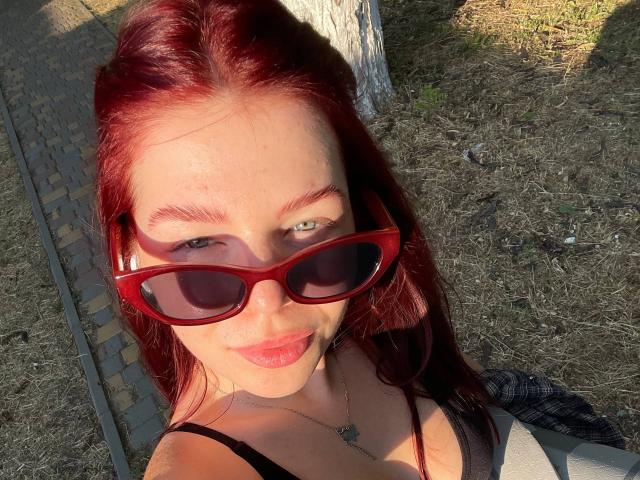 AlexXas, avec ses cheveux roux, portant des lunettes de soleil rouges, un haut noir et un collier, a les yeux verts et un léger sourire, et met en valeur sa poitrine et ses fesses dans la lumière du soleil..
