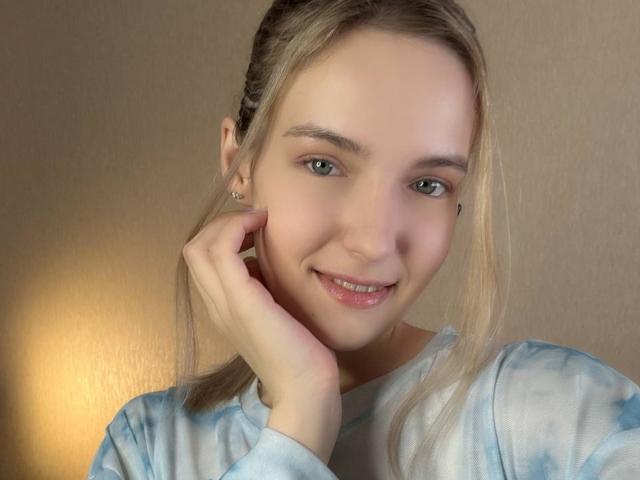 Nicole Spritzer ha i capelli biondi raccolti in trecce, la pelle chiara, gli occhi verdi e un sorriso carino. Indossa una maglietta colorata con motivi psichedelici e sta toccando la guancia con la mano destra..