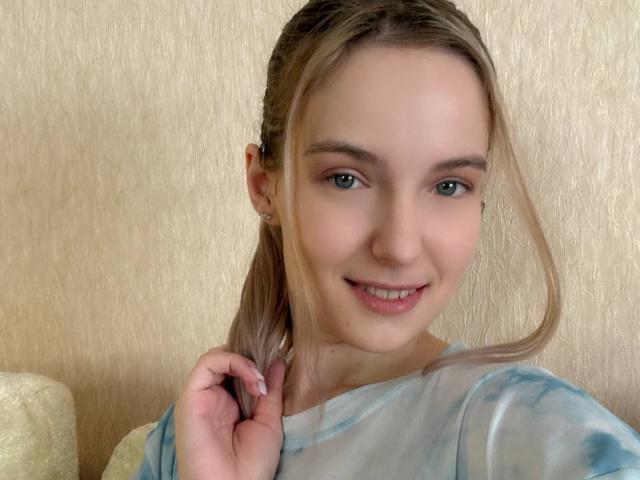NicoleSpritzer with blonde ponytail, green eyes, and slight smile, wearing a tie-dye shirt, holding her hair, showing her nose ring and earrings, against a beige textured wall.