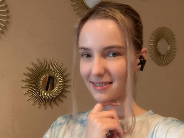 Nicole Spritzer mit blonden Haaren im Pferdeschwanz, lächelnd, mit einer Hand in der Nähe ihres Kinns, trägt ein blau-weißes T-Shirt im Batik-Stil und kleine schwarze Kopfhörer..