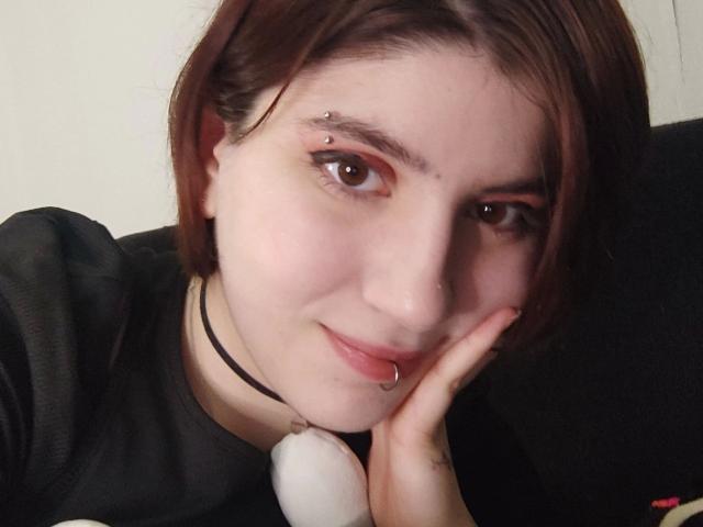 Julialooove-sexy, com cabelo castanho, olhos castanhos e um piercing no nariz, sorrindo para a câmera. Ela está vestindo uma camisa preta e segurando um brinquedo sexual branco na mão direita..
