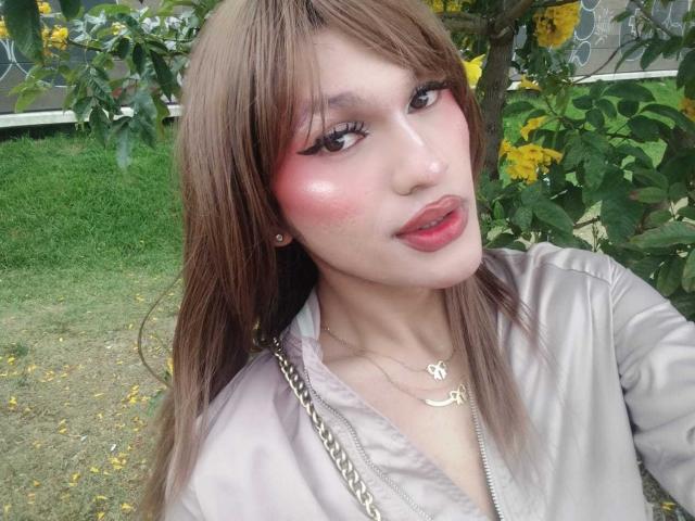 PamelaXFox tem bochechas rosadas e brilhantes, cabelos castanhos longos e veste uma jaqueta cinza brilhante. Seus lábios são vermelhos e ela está olhando para a câmera com um leve sorriso..