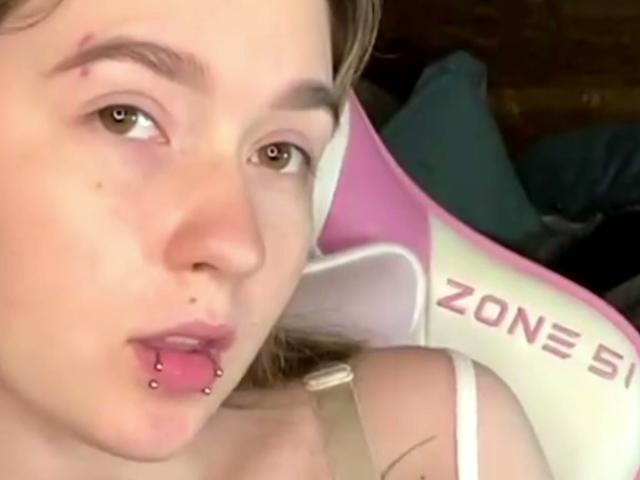 Liz Barrickman, con un piercing en la nariz, un piercing en el labio y piel clara, sentada en una silla de juego rosa y blanca, mirando a la cámara con la boca ligeramente abierta..