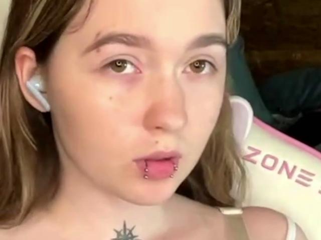 Liz Barrickman tiene una cara bonita con cabello castaño claro, ojos verdes, un piercing en la nariz y un piercing en el labio inferior. Lleva auriculares blancos y se puede ver una silla blanca..