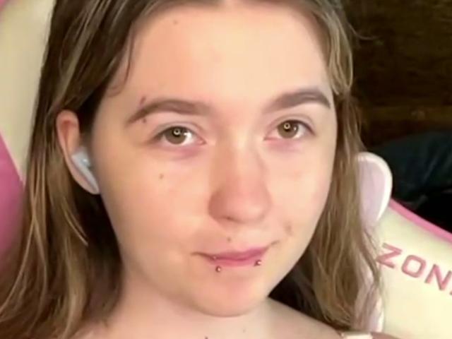 Liz Barrickman tem cabelo castanho claro, olhos verdes, um piercing no nariz, um piercing no lábio e uma pequena pinta na bochecha direita, e está usando fones de ouvido brancos..