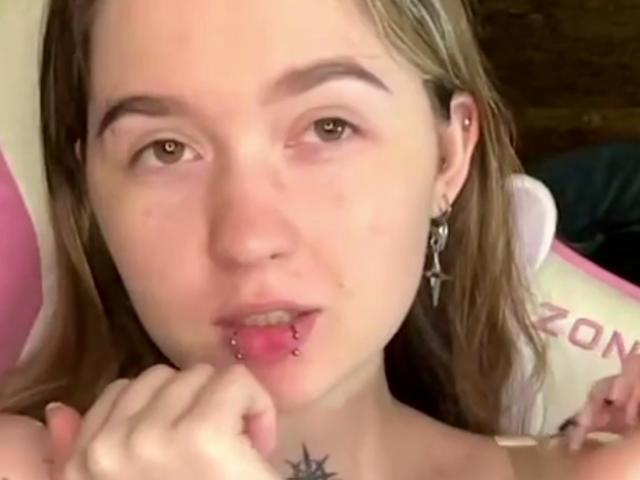 Liz Barrickman tem o lábio perfurado, exibindo joias cor-de-rosa, e tem uma mão perto da boca. Ela tem uma tatuagem de estrela abaixo do pescoço e usa um brinco de cruz..
