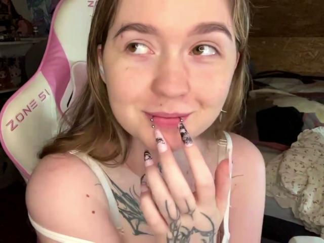 Liz Barrickman, com cabelo castanho claro, olhos verdes e batom rosa, exibindo suas unhas longas e pretas e sua mão tatuada, sorrindo levemente, vestindo uma camiseta branca sem mangas..