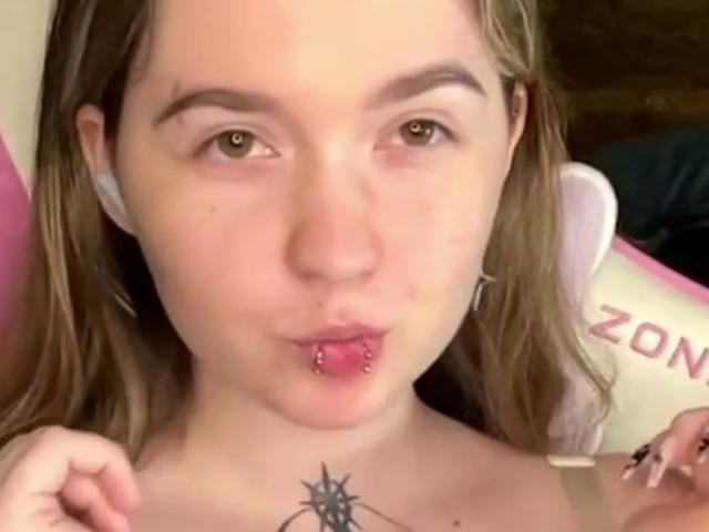 Liz Barrickman ha un labbro inferiore rosa con un piercing, capelli castano chiaro e un tatuaggio di una stella nera sotto il seno. Sta facendo una smorfia con le sue labbra rosa..