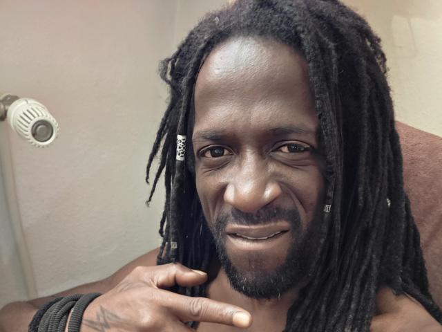 BigCazzoX, peau foncée, dreadlocks noirs de longueur moyenne, barbe de style bouc, souriant, main droite près du menton, portant des bracelets noirs, assis sur une chaise..