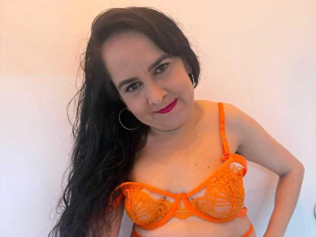 Diana Lane, avec de longs cheveux noirs, un soutien-gorge en dentelle orange, montrant ses seins, souriante, avec du rouge à lèvres rouge, des boucles d'oreilles cerceaux, debout devant un mur blanc, regardant l'appareil photo..