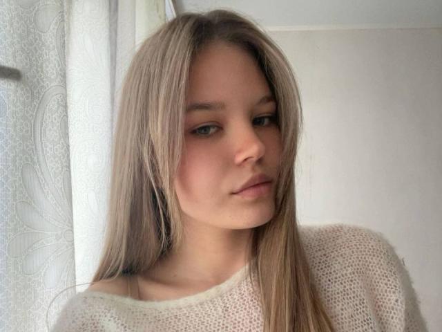 Theresa Hokett a de longs cheveux blonds et lisses, des yeux verts et une bouche légèrement retroussée. Elle porte un pull blanc en tricot. Son visage est légèrement tourné, ce qui révèle une expression détendue..