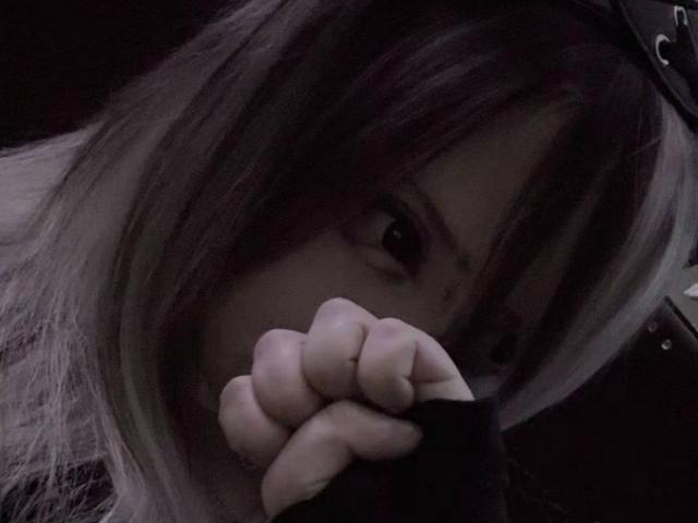 NovellaZellmer with dark, messy hair, eyes partially visible, clutching a black sleeve with her fist, showing a small scar under her right eye, in a dimly lit photo.