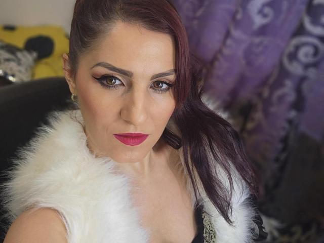 ShesTatiana, com cabelo ruivo, vestindo um colete branco e peludo, batom rosa e delineador escuro, olhando para cima com um leve sorriso, mostrando seus seios. Cortina roxa ao fundo..