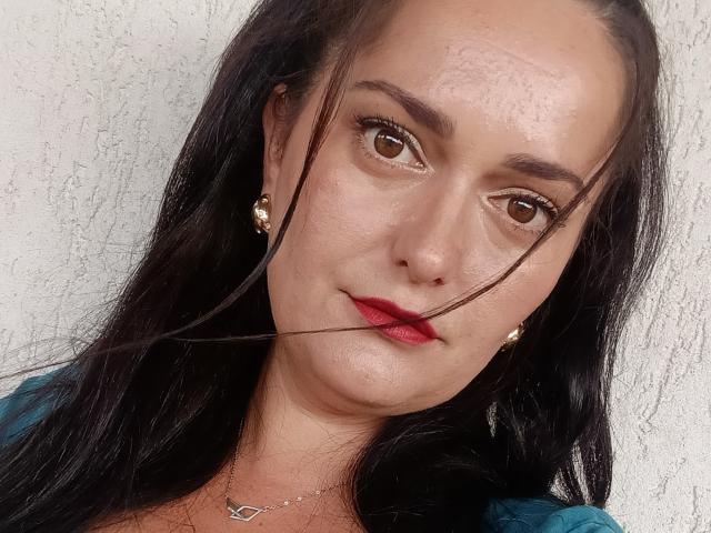 AmiraCeleste hat dunkles Haar, braune Augen, rote Lippen und eine leichte Haarsträhne im Mund. Sie trägt goldene Ohrringe und eine Halskette. Ihre Haut ist hell..