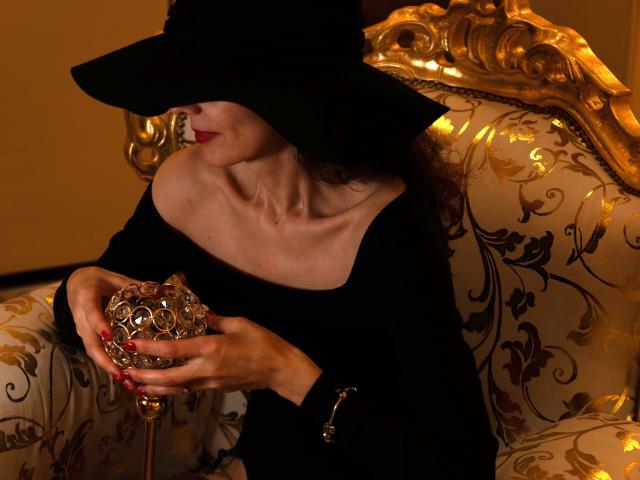 KamiLove com chapéu escuro, ombros descobertos, blusa preta, segurando uma bola dourada ornamentada, sentada em uma cadeira dourada e branca elegante, unhas vermelhas, iluminação marrom suave, clavícula visível..