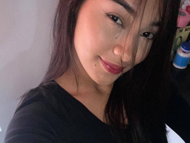 SashaHans with dark brown hair, light skin, red lipstick, small beauty mark under right eye, black shirt, slight smile, brown eyes, soft lighting, subtle makeup.