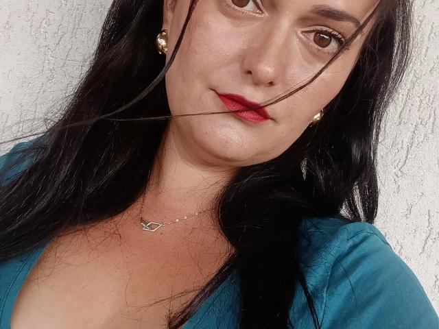 AmiraCeleste heeft lang zwart haar, bruine ogen, rode lippenstift en draagt een turquoise shirt met een ketting. Een enkele haar zit op haar lippen..