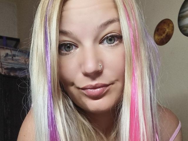 AlmaDeAngel69 ha i capelli biondi con riflessi rosa e viola, occhi azzurri, un piercing al naso e un leggero sorriso. Indossa una maglietta rosa e guarda direttamente la fotocamera..