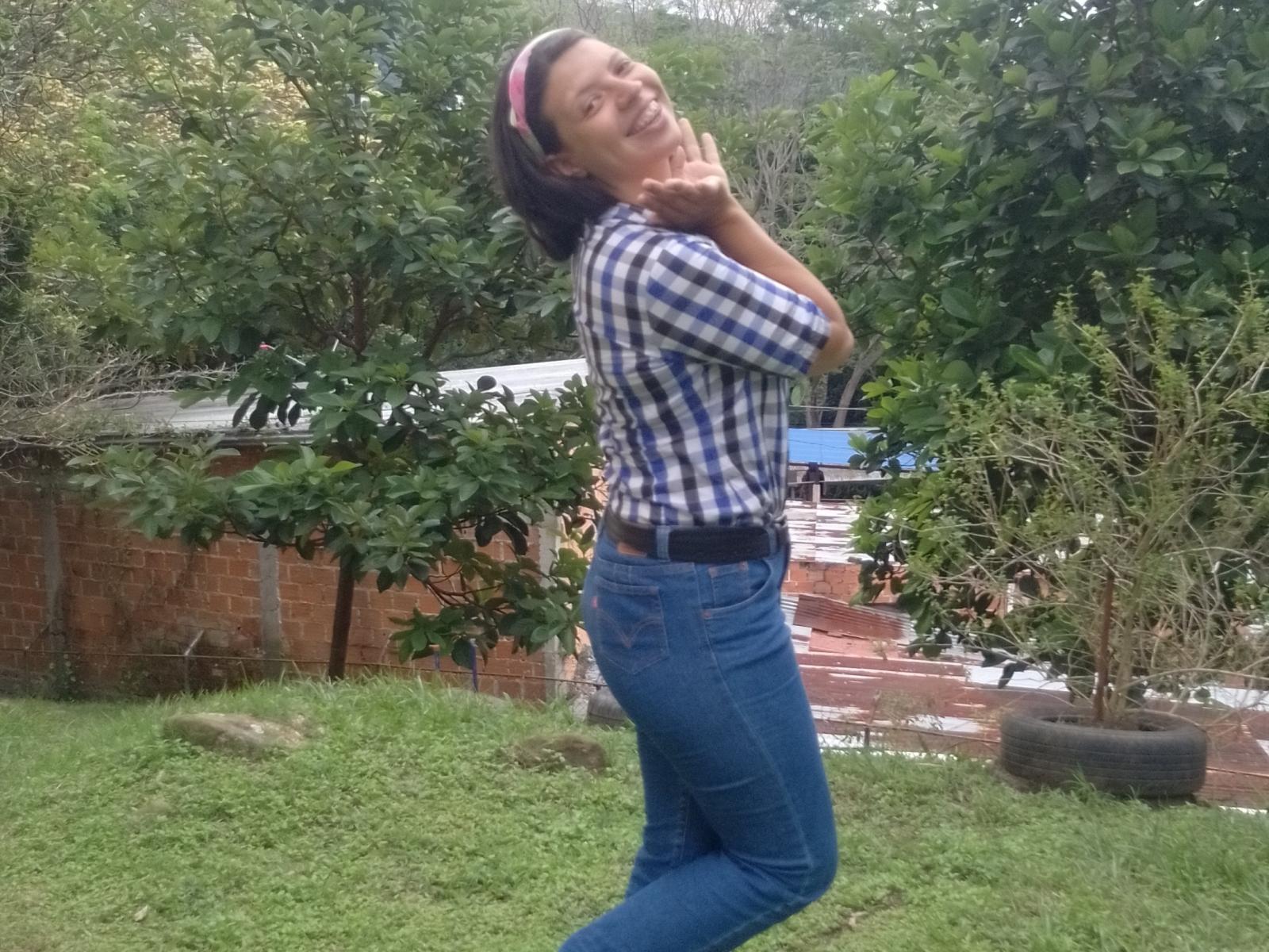 ConejitaHot69, con pechos de tamaño medio, vistiendo una camisa a cuadros azul y jeans azules, de pie sobre la hierba, con las manos cerca de la cara, sonriendo, con la cabeza inclinada, cabello castaño con una diadema rosa..