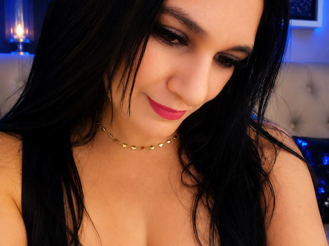 NicolleSmithh, con cabello negro, lápiz labial rosa y un collar dorado, mirando hacia abajo con una ligera sonrisa, mostrando sus senos. Lleva una blusa sin mangas, y su maquillaje es impecable..