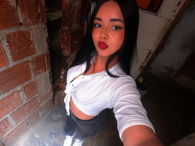 LunaSophiee mit langen schwarzen Haaren, rotem Lippenstift, einem weißen Crop-Top, schwarzen Hosen und weißen Sneakers, macht ein Selfie in einem Raum mit Ziegelwänden und zeigt dabei ihre Brüste..