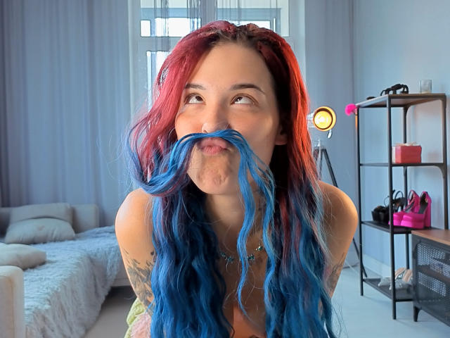 RachelNervis con capelli a sfumatura rossa e blu, la lingua fuori, che gioca con i suoi lunghi baffi blu, con un piercing al naso e tatuaggi visibili, in una stanza luminosa con un treppiede e una mensola..