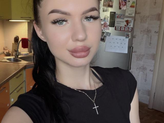 DaineKately, con la pelle chiara, gli occhi azzurri, i capelli neri raccolti in una coda di cavallo, indossa una maglietta nera e una croce. Ha labbra piene di colore rosa e lunghe ciglia. Sullo sfondo si vede una cucina..