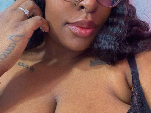 NatyLm's face with dark wavy hair, wearing glasses, touching her cheek, showing tattoos, and black lace bra, exposing her tits, with a ring on her finger, and pink lips.