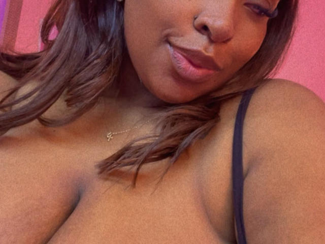 NatyLm's brown face, pouty pink lips, and wavy dark brown hair, wearing a black strap, with a gold necklace and visible cleavage, smiling slightly.